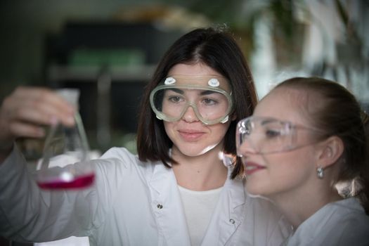 Chemical laboratory. Two young woman holding a flask with pink liquid in it, smiling. Close up