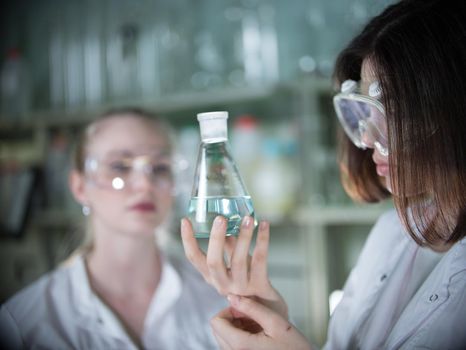 Two young woman in chemical lab holding a flasks with clear liquid in it. Flask in focus