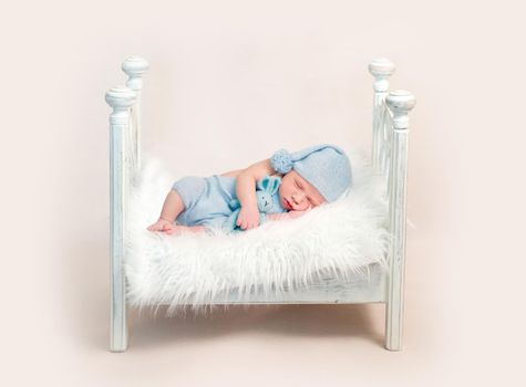 Newborn baby boy sleeping on white wooden crib embracing hare toy. Little boy wearing blue outfit sleeps on small bed