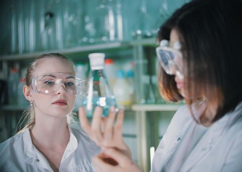 Two young woman in chemical lab holding a flasks with clear liquid in it. Blonde woman in focus. Portrait