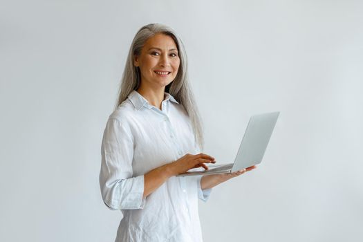 Attractive mature Asian business lady with grey hair in elegant blouse holds open modern laptop on light background in studio