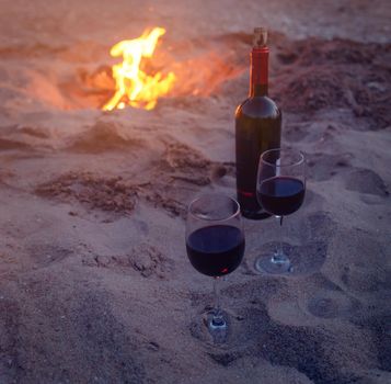 Bottle and two glasses with red wine on sand beach near campfire, concept of romantic date.