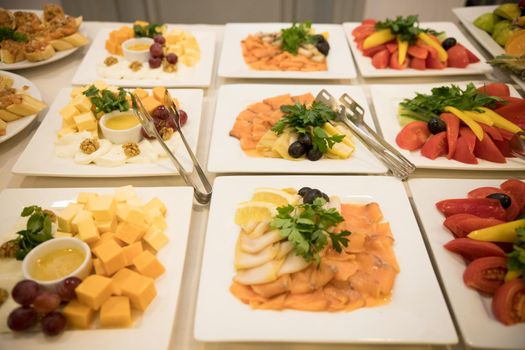 Row of plates with various cold appetizers standing on a table with a white tablecloth. Close up