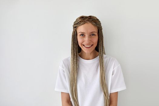 Cheerful attractive female with braids wearing casual white shirt standing on light background and looking at camera