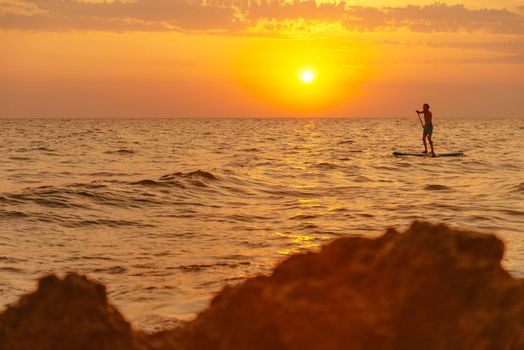 Young man floating on a paddle board, SUP, in the sea at sunset, rear view.