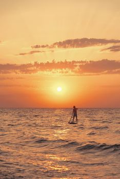 Silhouette of young man floating on stand up paddle board, SUP, in the sea at sunset.