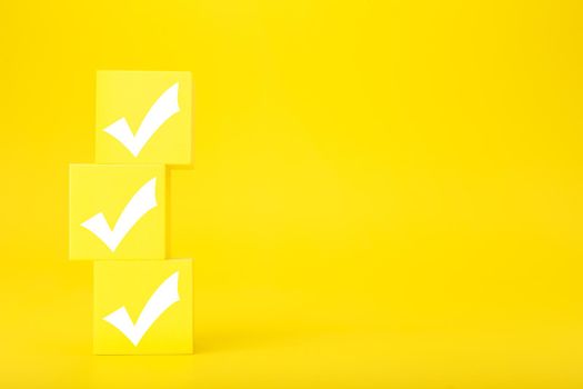 Three white checkmarks on stack of yellow toy cubes on bright yellow background with copy space. Concept of questionary, kids related checklist, to do list, planning, business or verification.