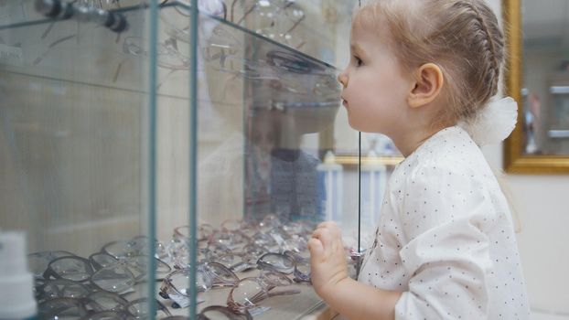 Adorable child blonde girl in ophthalmology clinic plays in hall near glasses store, close up