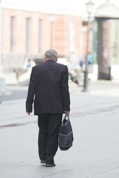 An old man in a black suit walks down the street with a tote bag in his hands. Wide shot
