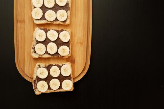 Three banana white bread toasts spread with chocolate butter that lie on a chopping board on a dark background. top view with area for text
