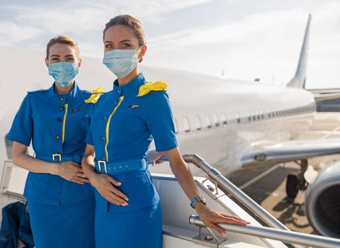 Two beautiful air stewardesses in blue uniform and protective face masks looking at camera, standing on airstair during boarding. Aircrew, occupation, covid19 concept
