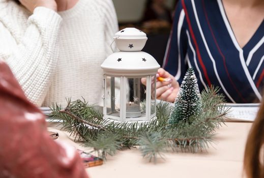 A white metal candlestick with candle inside surrounded with christmas tree. Women sitting around the table. Christmas party or meeting.