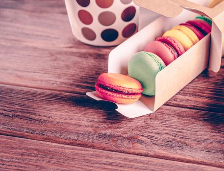 Delicious sweet macaroons in a box near the cup on a wooden table.