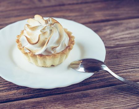 Cream cupcake on a white saucer with teaspoon on a wooden table.