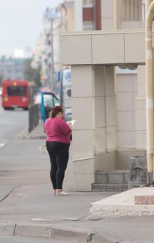 Plus-size young woman stands back to the camera and writes in a notebook on the street. Wide shot