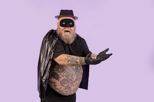 Emotional plus size man in black hero suit with large bare tummy shows on something standing on purple background in studio