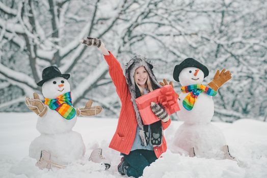 Girl playing with snowman in winter park. Snowman and funny young woman in winter hat and scarf