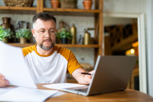 A middle-aged man works with papers at home in front of a laptop monitor. He is sitting with glasses and typing text. Modern technology concept.