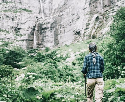 Explorer young woman with trekking poles walking to waterfalls on high cliff.