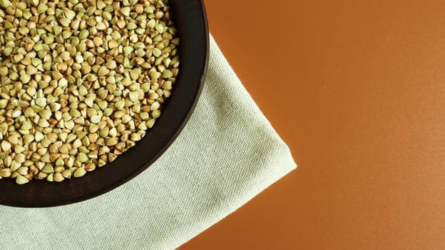 Raw green buckwheat in a brown clay plate on a brown background. Vegan organic food concept. The concept of diet, weight loss, healthy and proper nutrition. Copy space for text. Top view, flat lay.