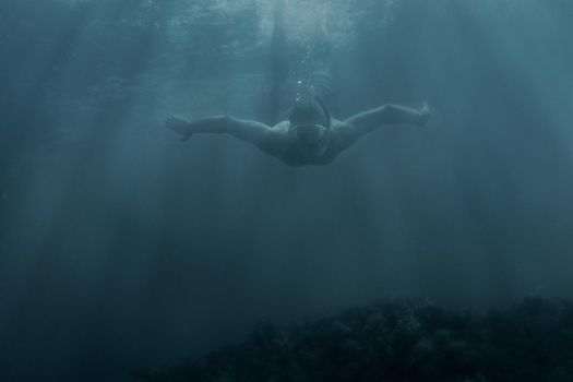 Active male freediver in mask swimming underwater among sunbeams, front view.