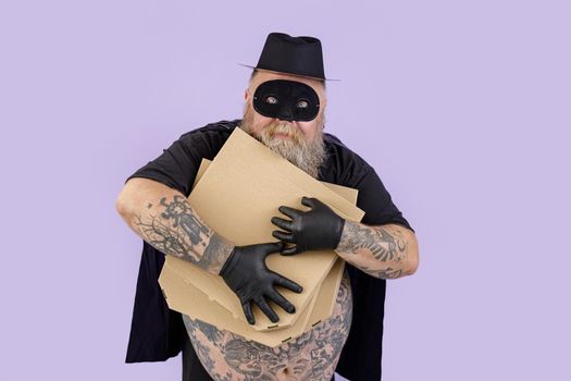 Greedy mature man in Zorro costume with overweight and tattoos hugs brown boxes of pizza on purple background in studio