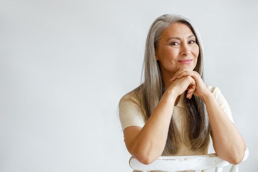 Happy middle aged Asian woman with loose silver hair sits on chair leaning on hands on light background in studio. Mature beauty lifestyle