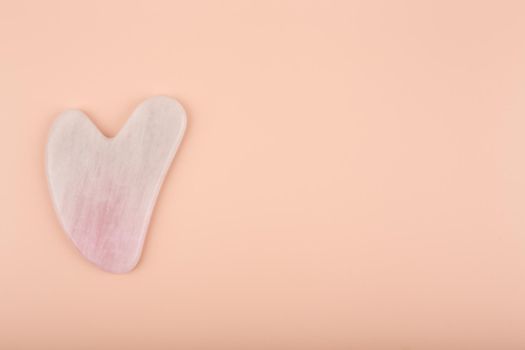 Top view of pink heart shaped guasha stone made of quartz crystal on bright beige background with copy space. Concept of alternative skin care treatment, self massage and acupressure