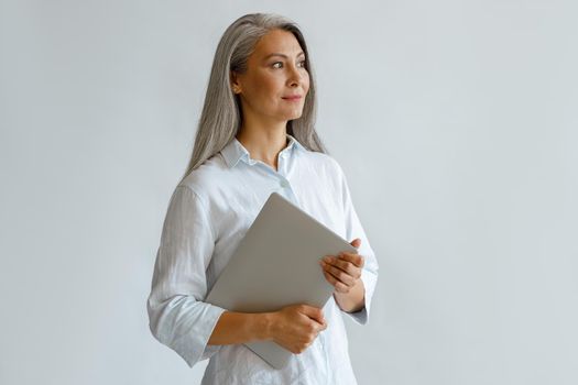 Positive mature Asian businesswoman wearing white blouse holds contemporary laptop standing on light grey background in studio