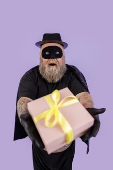 Excited mature bearded gentleman with overweight wearing carnval Zorro costume with cape and mask presents gift on purple background in studio