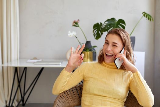 Happy woman in yellow turtleneck winking and showing okay gesture while sitting on chair and talking on mobile phone in light workspace