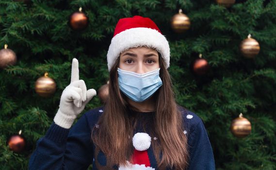 Christmas idea.Close up Portrait of woman wearing a santa claus hat and medical mask with emotion. Against the background of a Christmas tree. Coronavirus pandemic