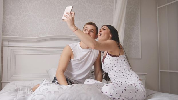 Happy cheerful loving couple making selfie in bed, young attractive guy and girl sitting in pajamas, people's attitudes