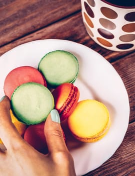 Female hand takes sweet macaroon cookie near the cup on wooden table, top view.
