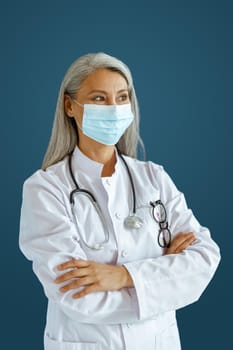 Hoary haired Asian woman doctor in uniform with surgical mask and crossed arms stands on blue background in studio. Professional medical staff