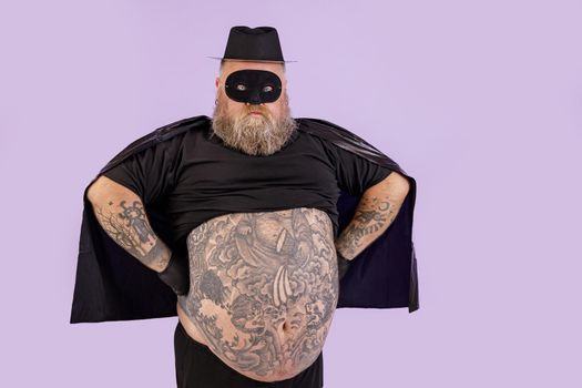 Confident mature man with overweight wearing Zorro suit with cape and mask holds hands on waist standing on purple background in studio