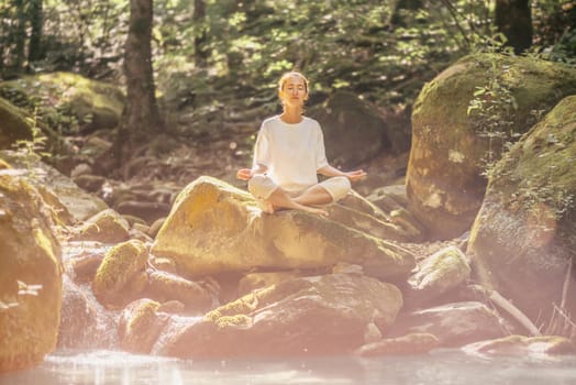 Beautiful young woman practicing yoga with closed eyes in pose of lotus on stone near the river in forest outdoor.