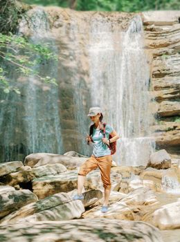 Explorer backpacker young woman walking on stones in front of waterfall outdoor on sunny day.