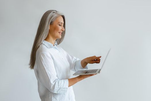 Smiling grey haired mature Asian businesswoman in white blouse points onto screen of modern laptop on light background in studio side view