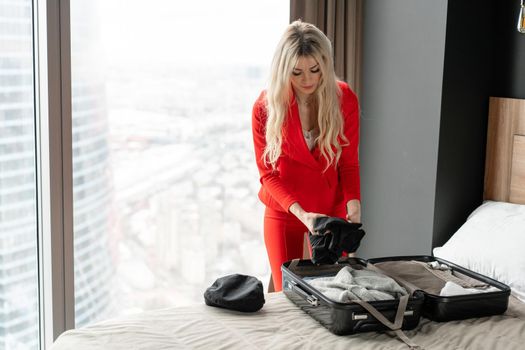 Stewardess in red uniform arrives in a hotel room with black suitcase. Rest in the transit city before the return flight. Young blond Woman in red coral suit unpacks suitcase