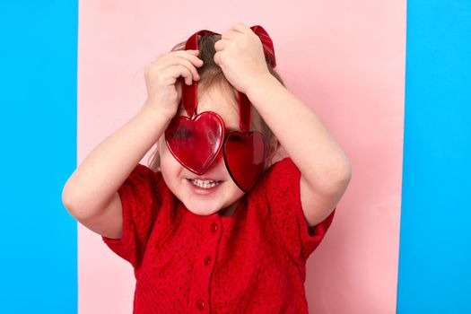 Cute delighted little girl standing in studio and looking at camera through red plastic toy hearts