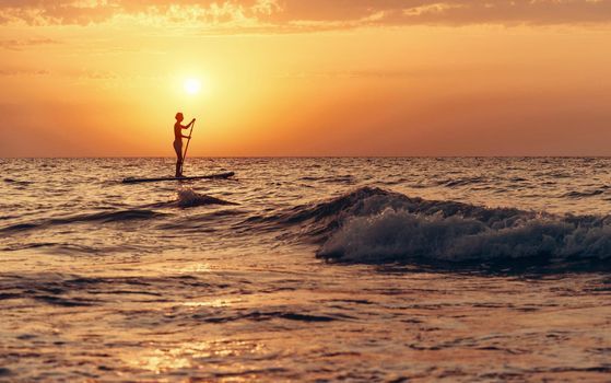 Silhouette of young man paddling on a SUP board in the sea at sunset.