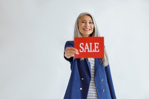 Beautiful mature Asian woman with loose grey hair wearing blue jacket shows red card with word Sale posing on light background in studio, space for text