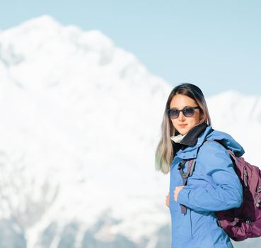 Backpacker young woman standing on background of snowy mountains, looking at camera.