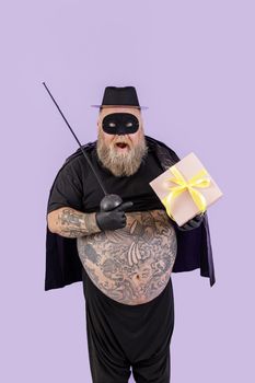 Surprised mature man with overweight in Zorro suit with toy epee points onto gift box with yellow bow on purple background in studio