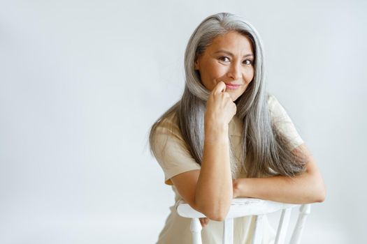Flirting middle aged Asian lady with beautiful grey hair sits on chair on light background in studio. Mature beauty lifestyle