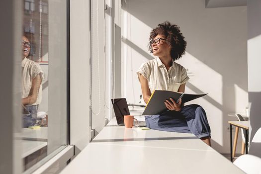 Happy multiethnic woman in glasses sitting on the table while holding notebook and looking out the window