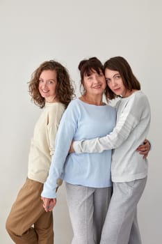 Cheerful adult mother embracing charming young daughters while standing on white background in studio and looking at camera