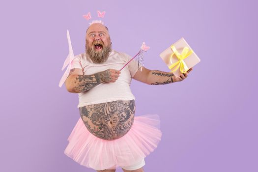 Excited bearded obese male person in fairy costume with magic stick and wings holds gift box on purple background in studio