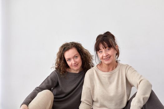 Cheerful adult daughter and middle aged mother sitting on floor at home on white background